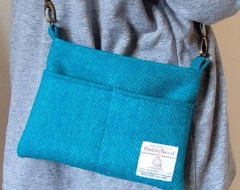 May include: A teal blue tweed crossbody bag with a brown leather strap and a small label that reads "Harris Tweed" with a small logo of a ball of yarn with a cross through it. The bag is worn by a person wearing a gray sweater and black pants.
