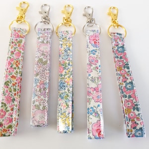 May include: Five colourful floral patterned keychains with gold and silver key rings. The keychains are made of fabric and have a white background with pink, blue and yellow flowers.