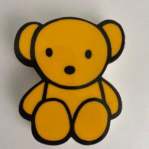 Puede incluir: Un oso de dibujos animados amarillo con contornos negros, sentado con las piernas cruzadas. El oso tiene una cara simple con ojos, nariz y boca negros.