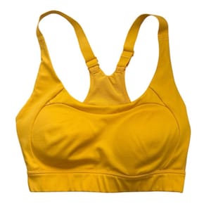 Puede incluir: Sujetador deportivo amarillo mostaza con tirantes ajustables y diseño de espalda deportiva. El sujetador presenta copas moldeadas y una banda ancha debajo del busto para brindar soporte. La tela parece ser un material liso y que absorbe la humedad, adecuado para actividades atléticas.