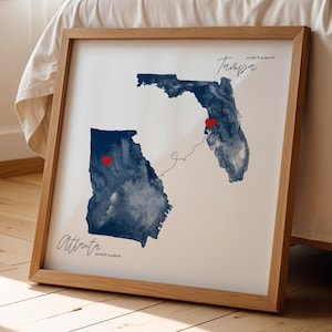 May include: A square framed wall art print featuring watercolor-style navy blue silhouettes of Georgia and Florida. Two red hearts mark locations connected by a thin line. The text reads Atlanta and Tampa with coordinates in black script.