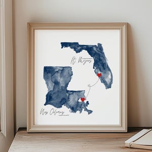 May include: A framed watercolor print featuring dark blue watercolor maps of Louisiana and Florida, with red heart accents. The cities of New Orleans and Fort Myers are labeled in cursive script. The print is displayed on a wooden table.