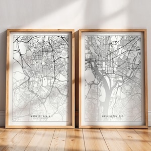 Op de afbeelding: Twee ingelijste prints van stadskaarten. Eén toont Madrid, Spanje, en de andere Washington, D.C. De kaarten zijn in zwart-wit, met gedetailleerde straatindelingen. De frames zijn van lichtgekleurd hout en de prints worden op een houten vloer getoond.