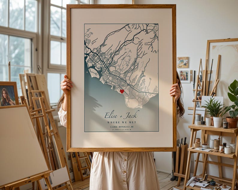 May include: A framed map print featuring a detailed city map with a heart marking a specific location. The map is in shades of blue and beige, with the names "Elise + Jack" and the phrase "WHERE WE MET" displayed below the map.