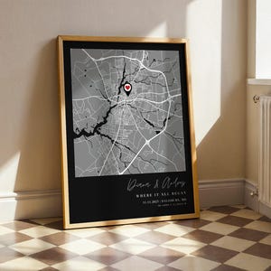 May include: Framed map print with a black and white city map design. The map features a red heart marker and the text "Where it all began" with a date and location. The frame is a light wood color.