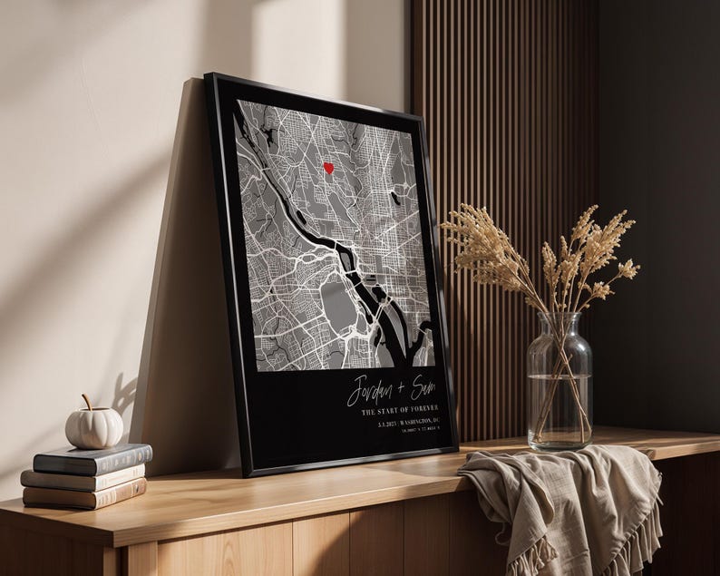 May include: Framed black and white map print with a red heart marking a location. The print includes the names "Jordan + Sam" and the text "THE START OF FOREVER" with a date and location. The print is displayed on a wooden surface.