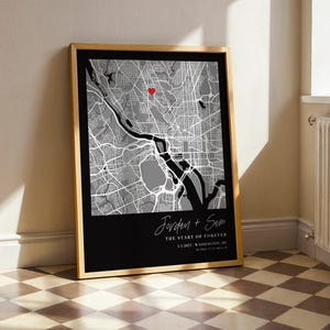 May include: Framed map art print featuring a grayscale city map with a red heart marker. The bottom section is black with white text that reads "Jordan + Sam" and "The Start of Forever". The frame is a light wood color.