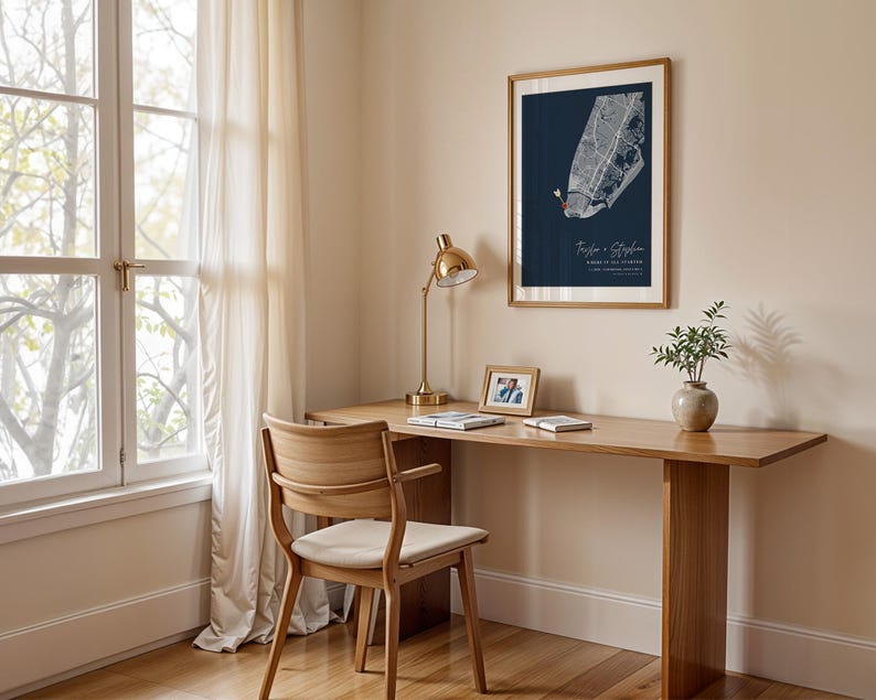 May include: A framed map print with a navy blue background hangs on a beige wall. The map is white with red accents. The text "Taylor & Stephen" is below the map. A wooden desk with a gold lamp, a small photo frame, and a vase with greenery sits below the print.