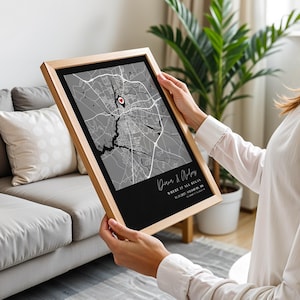 May include: A framed map print with a black and gray color scheme. The map features a red heart-shaped marker. The frame is made of light-colored wood. The print includes text at the bottom, with the words "Where it all began."