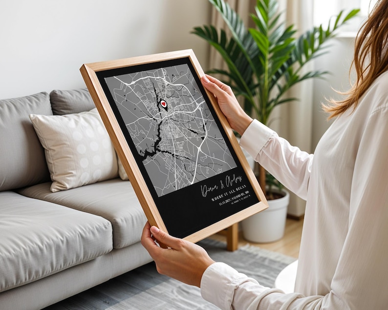 May include: A framed map print with a black and gray color scheme. The map features a red heart-shaped marker. The frame is made of light-colored wood. The print includes text at the bottom, with the words "Where it all began."