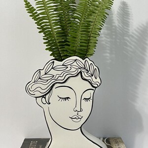 May include: A white ceramic face-shaped vase with a black outline, holding green fern leaves. The vase is decorated with a laurel wreath and closed eyes. The vase is sitting on top of a book titled "BLACK & WHITE (and a bit in between)".