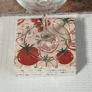 May include: A stack of square coasters with a tomato design. The coasters feature a repeating pattern of red tomatoes and green leaves on a beige background. A clear wine glass sits on top of the coasters.