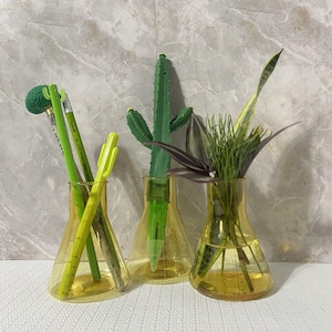 May include: Three yellow glass beakers with various items inside. One beaker holds green pens, another a cactus pen, and the third has plants. The beakers are on a white surface with a marble-patterned background.