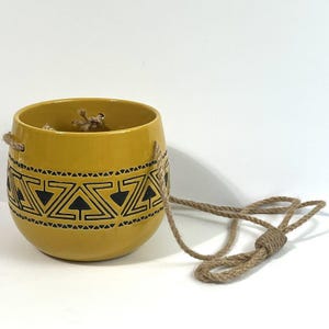 May include: Mustard yellow ceramic hanging planter with a black geometric design. The planter has a rope for hanging. The planter is round with a wide opening. The planter is designed for indoor or outdoor use.