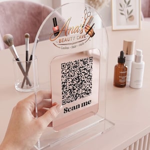 May include: Clear acrylic sign for a beauty salon, featuring the text "Ana's Beauty Cave" with a logo of nail polish, scissors, and a comb. A QR code is displayed below the logo, with the words "Scan me".