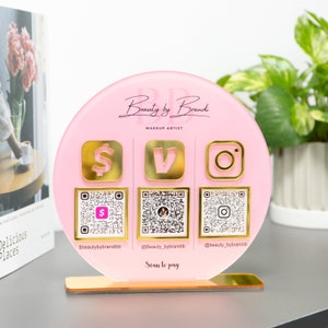 May include: A pink round acrylic sign with gold accents featuring the text "Beauty by Brandi Makeup Artist". The sign has three gold icons: a dollar sign, a letter V, and an Instagram logo. Each icon has a QR code underneath it. The sign is on a gold stand.