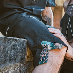 Può includere: Una coppia vestita di nero con accenti colorati. L'uomo indossa una camicia nera abbottonata e pantaloncini neri con un bordo a motivi colorati. La donna indossa un top nero con una sovrapposizione grigia e pantaloni neri con accenti argentati. Entrambi indossano occhiali da sole.