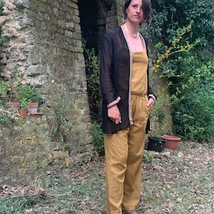 Può includere: Una donna indossa un cardigan nero trasparente sopra una tuta gialla senape. È in piedi davanti a un muro di pietra con un arco.