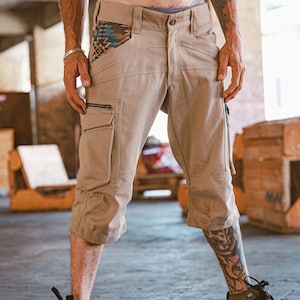 Peut inclure: Short cargo beige avec un détail de poche à motif. Le short a une poche zippée sur la jambe gauche et une poche à rabat sur la jambe droite. Le short est porté par une personne avec des tatouages sur les jambes.