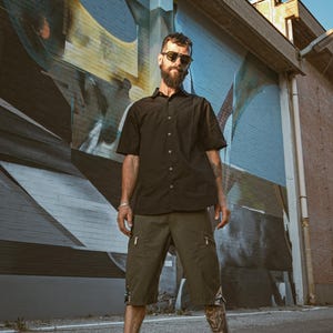 Peut inclure: Un homme portant une chemise boutonnée noire, un short cargo vert foncé et des lunettes de soleil noires se tient devant un mur de graffitis. Il porte des chaussures noires et a une barbe.
