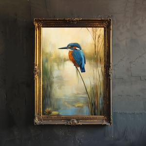 Puede incluir: Pintura enmarcada de un martín pescador posado en una caña. El ave, con plumaje azul y naranja, se alza sobre un paisaje lacustre sereno con juncos y nenúfares. El marco dorado ornamentado añade un toque clásico a la obra.