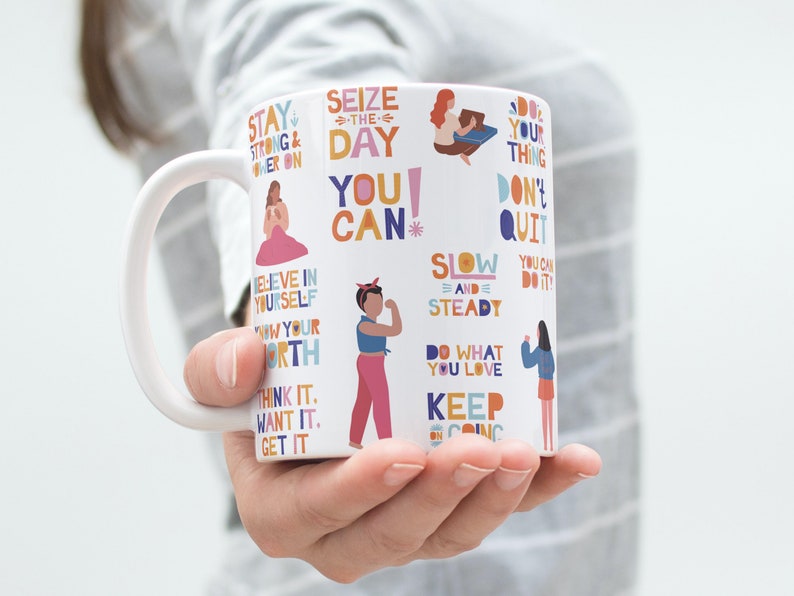 Peut inclure: Une tasse en c&eacute;ramique blanche avec du texte et des illustrations motivants color&eacute;s. Le texte comprend des phrases comme "Saisissez le jour", "Vous pouvez", "Lentement et s&ucirc;rement", "Faites ce que vous aimez", et "Continuez".