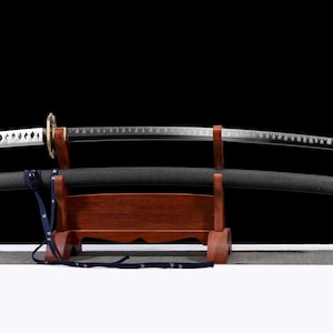 Puede incluir: Una espada katana con una vaina negra y una hoja plateada descansa sobre un soporte de madera. El mango es blanco con detalles en forma de diamante. La guarda de la espada es dorada y el soporte es de color marr&oacute;n oscuro.