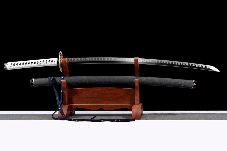 Puede incluir: Una espada katana con una vaina negra y una hoja plateada descansa sobre un soporte de madera. El mango es blanco con detalles en forma de diamante. La guarda de la espada es dorada y el soporte es de color marr&oacute;n oscuro.