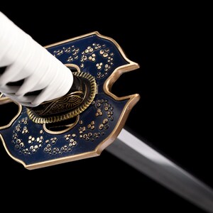 Puede incluir: Primer plano de una katana. El mango est&aacute; envuelto en tela blanca, y la guarda es azul oscuro con detalles dorados y motivos florales. La hoja es plateada e inclinada, sobre un fondo negro.