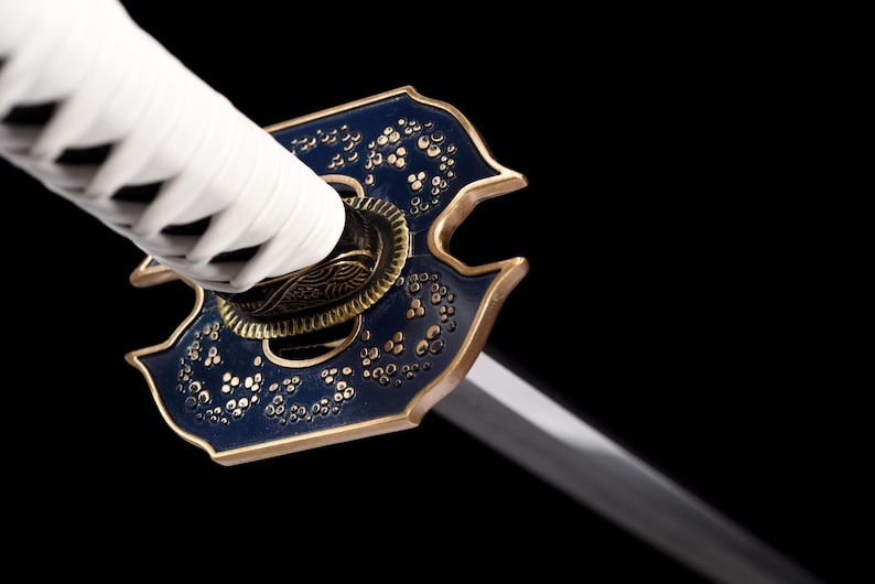 Puede incluir: Primer plano de una katana. El mango est&aacute; envuelto en tela blanca, y la guarda es azul oscuro con detalles dorados y motivos florales. La hoja es plateada e inclinada, sobre un fondo negro.