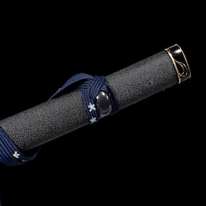 Puede incluir: Primer plano de una empu&ntilde;adura de espada negra con una superficie texturizada. Un cord&oacute;n azul marino con detalles florales blancos est&aacute; envuelto alrededor de la empu&ntilde;adura. La tapa del extremo es negra con ribete dorado.