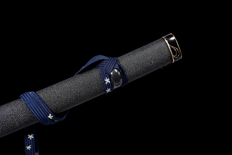 Puede incluir: Primer plano de una empu&ntilde;adura de espada negra con una superficie texturizada. Un cord&oacute;n azul marino con detalles florales blancos est&aacute; envuelto alrededor de la empu&ntilde;adura. La tapa del extremo es negra con ribete dorado.