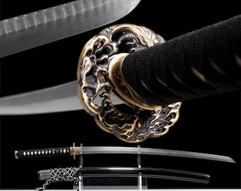 May include: Close-up of a Japanese katana sword with a black handle and a decorative gold and bronze tsuba. The blade is silver with a visible pattern. A second katana is displayed below, sheathed in a black scabbard.