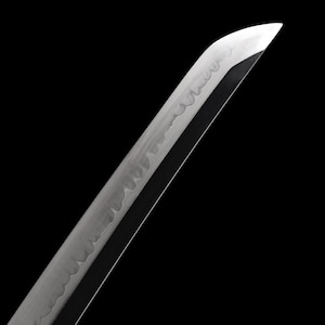 Puede incluir: Primer plano de una hoja de katana sobre un fondo negro. La hoja es plateada con un filo afilado y un patr&oacute;n visible a lo largo del lateral. La punta de la hoja est&aacute; angulada, y el dise&ntilde;o general sugiere una espada japonesa tradicional.
