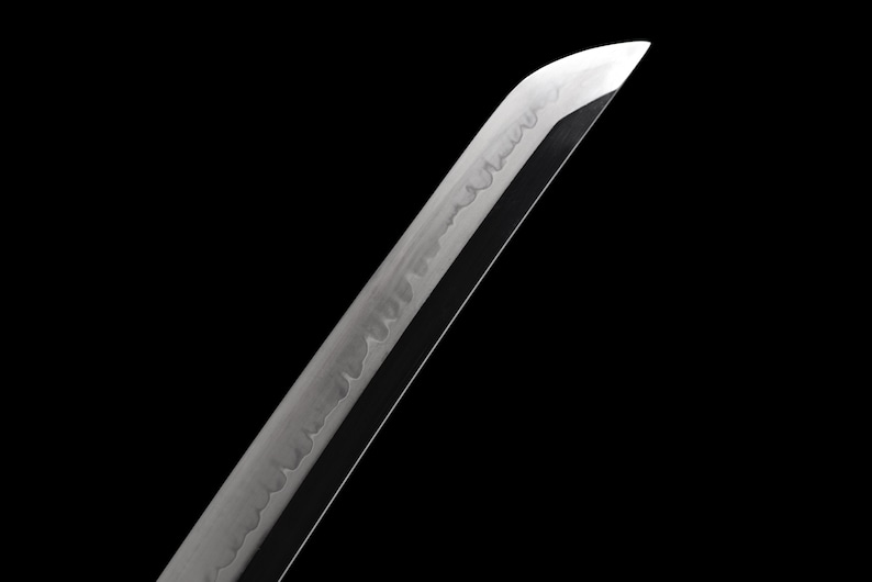 Puede incluir: Primer plano de una hoja de katana sobre un fondo negro. La hoja es plateada con un filo afilado y un patr&oacute;n visible a lo largo del lateral. La punta de la hoja est&aacute; angulada, y el dise&ntilde;o general sugiere una espada japonesa tradicional.