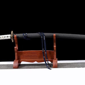 Puede incluir: Una katana japonesa con vaina negra y empu&ntilde;adura blanca, apoyada en un soporte de madera. La empu&ntilde;adura presenta un patr&oacute;n de diamantes y una correa azul con estrellas blancas cuelga de la vaina. La espada se exhibe sobre un fondo negro.