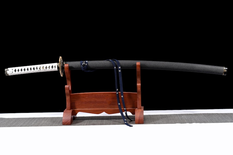 Puede incluir: Una katana japonesa con vaina negra y empu&ntilde;adura blanca, apoyada en un soporte de madera. La empu&ntilde;adura presenta un patr&oacute;n de diamantes y una correa azul con estrellas blancas cuelga de la vaina. La espada se exhibe sobre un fondo negro.
