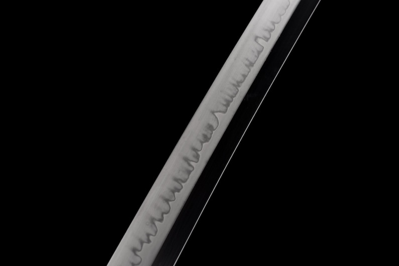 Puede incluir: Primer plano de una hoja de katana con un patr&oacute;n detallado a lo largo del filo. La hoja tiene un acabado plateado y est&aacute; sobre un fondo negro. El dise&ntilde;o de la hoja es visible.
