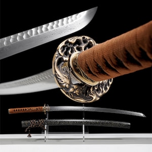 Puede incluir: Una espada katana japonesa con un mango marrón y una guarda decorativa dorada. La hoja es plateada con un patrón detallado. La espada se exhibe sobre un soporte transparente, con su vaina negra debajo.