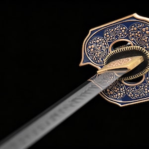 Puede incluir: Primer plano de una katana japonesa. La guarda decorativa azul y dorada presenta un intrincado patr&oacute;n floral. La hoja es plateada con detalles grabados y el mango est&aacute; envuelto en tela blanca. El fondo es negro.