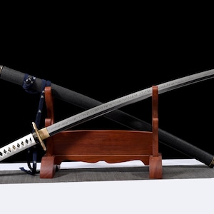 Puede incluir: Una katana japonesa con empu&ntilde;adura blanca y hoja plateada, apoyada en un soporte de madera. La vaina negra est&aacute; colocada detr&aacute;s de la hoja. La espada se exhibe sobre un fondo negro.