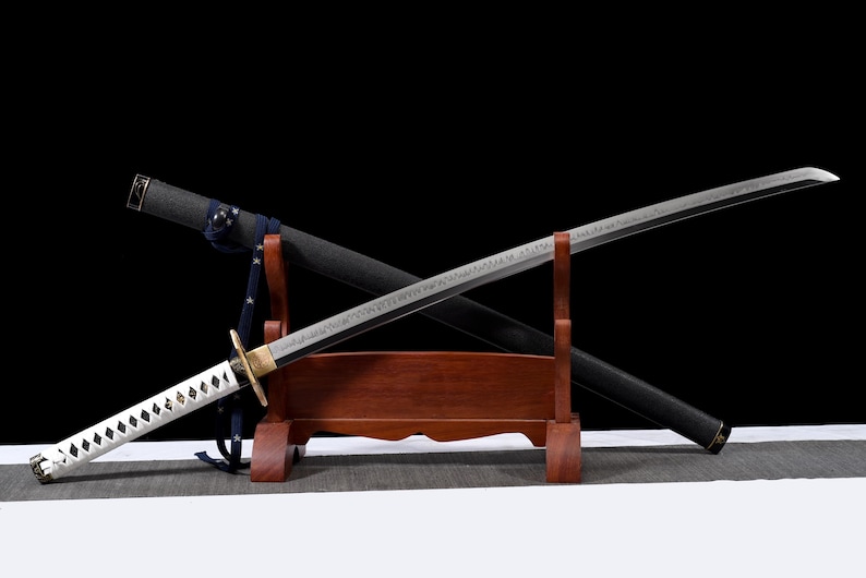 Puede incluir: Una katana japonesa con empu&ntilde;adura blanca y hoja plateada, apoyada en un soporte de madera. La vaina negra est&aacute; colocada detr&aacute;s de la hoja. La espada se exhibe sobre un fondo negro.