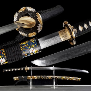 Puede incluir: Una espada katana japonesa con mango envuelto en negro, una guarda floral en oro y plata, y una hoja con una línea de temple ondulada visible. La vaina presenta un patrón moteado colorido y detalles florales blancos, exhibida sobre un soporte.