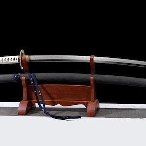 Puede incluir: Una espada katana japonesa con una hoja curva, expuesta sobre un soporte de madera. La espada presenta un mango blanco con un dise&ntilde;o de diamantes y una vaina negra. Una correa azul cuelga de la vaina. El fondo es negro.