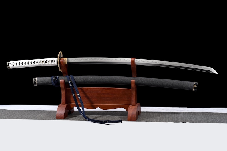 Puede incluir: Una espada katana japonesa con una hoja curva, expuesta sobre un soporte de madera. La espada presenta un mango blanco con un dise&ntilde;o de diamantes y una vaina negra. Una correa azul cuelga de la vaina. El fondo es negro.