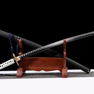 Puede incluir: Una espada katana japonesa con una vaina negra y un mango blanco y negro. La hoja es plateada con un borde decorativo. La espada descansa sobre un soporte de madera con una correa azul.