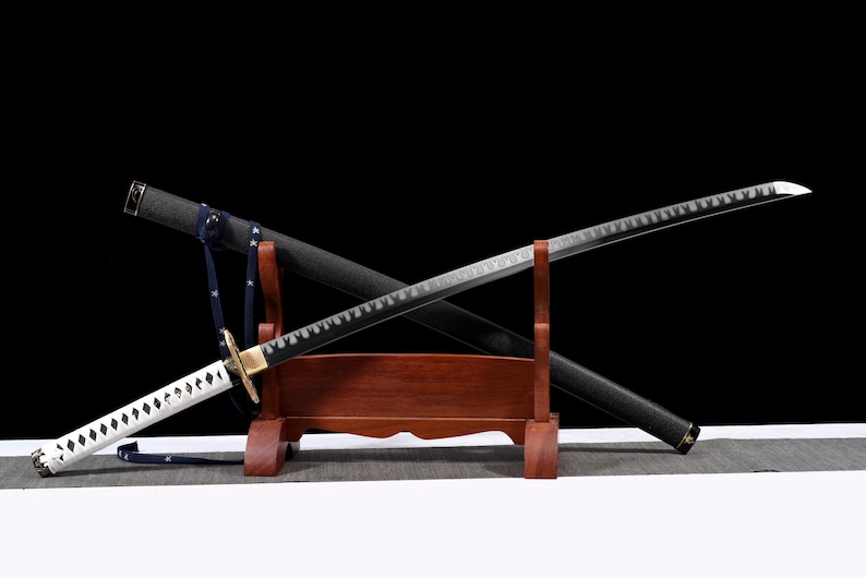 Puede incluir: Una espada katana japonesa con una vaina negra y un mango blanco y negro. La hoja es plateada con un borde decorativo. La espada descansa sobre un soporte de madera con una correa azul.