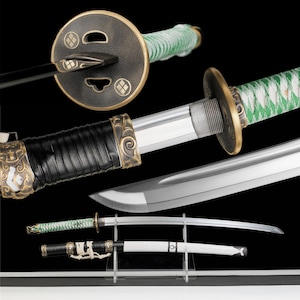 Op de afbeelding: Een katana met een groen omwikkeld handvat en een ronde, bronskleurige stootplaat met een textuurpatroon. Het lemmet is van zilverkleurig staal. Een aparte witte schede met zwarte accenten en een zwart koord ligt op een doorzichtige standaard.