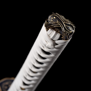 Puede incluir: Primer plano del mango de una katana. El mango est&aacute; envuelto en tela blanca con un pomo decorativo negro y dorado. El pomo presenta intrincados dise&ntilde;os dorados sobre un fondo negro. La hoja no es visible.