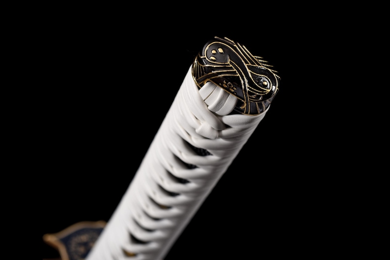 Puede incluir: Primer plano del mango de una katana. El mango est&aacute; envuelto en tela blanca con un pomo decorativo negro y dorado. El pomo presenta intrincados dise&ntilde;os dorados sobre un fondo negro. La hoja no es visible.